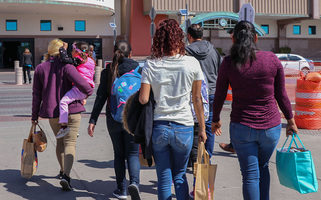 Un grupo de personas con bolsas de compras caminando hacia un gran edificio en un día soleado.