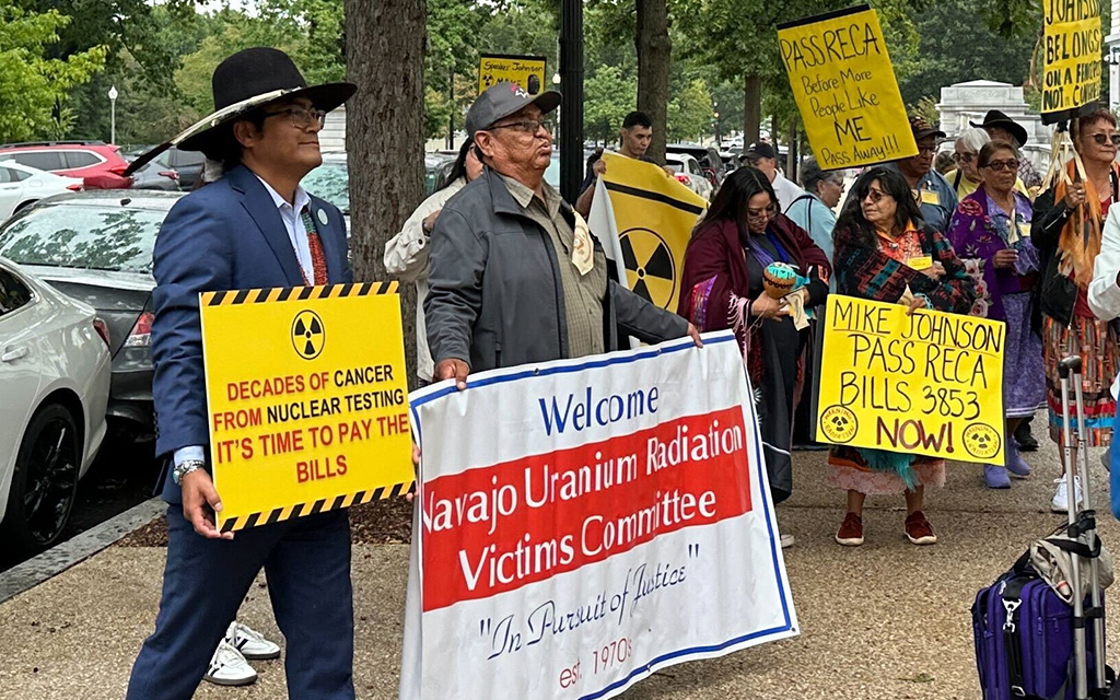 Group of people holding signs during a protest about the effects of nuclear testing, with a prominent banner reading "Welcome Navajo Uranium Radiation Victims Committee."