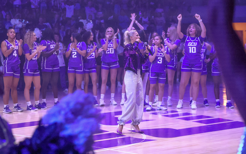 GCU women’s basketball coach Molly Miller with a microphone stands on a basketball court surrounded by the GCU female basketball players in purple uniforms.