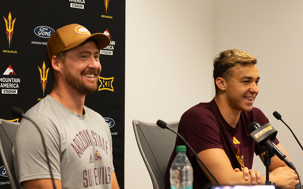ASU swimmer Ilya Kharun on the right and another individual on the left at a press conference table with microphones and a backdrop featuring various logos.