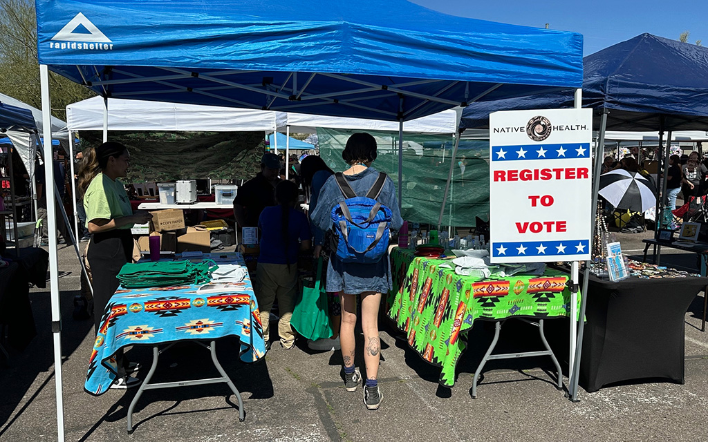 Outdoor voter registration booth with "REGISTER TO VOTE" sign under a blue canopy.