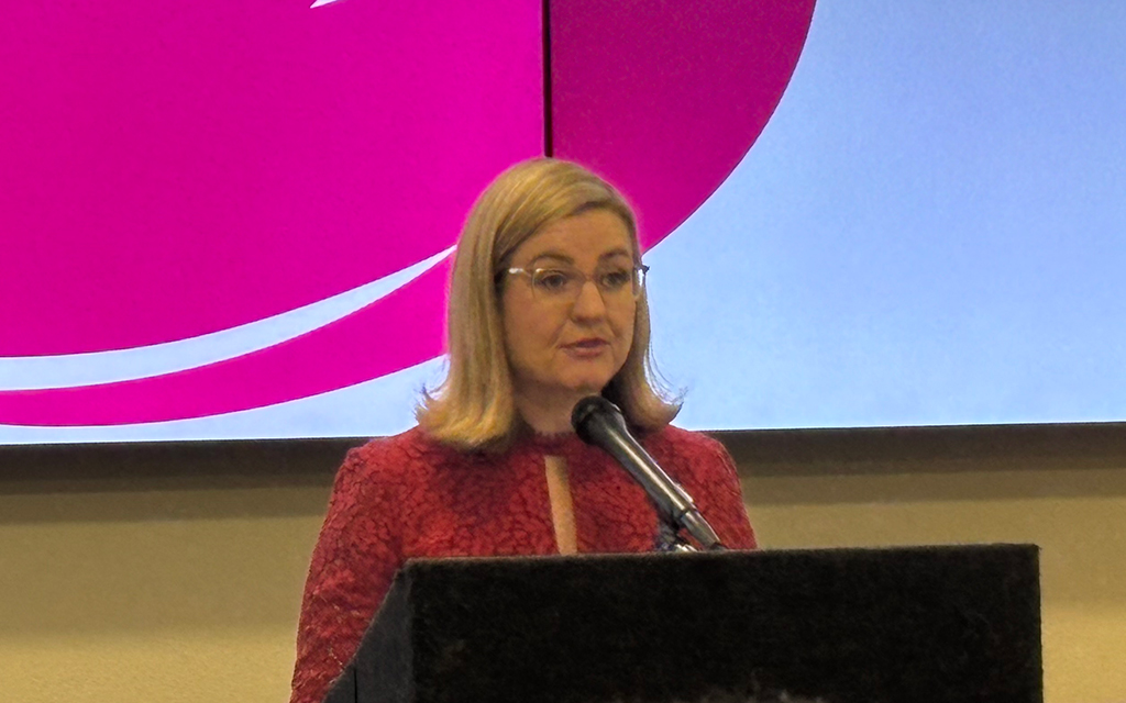 Kate Gallego speaking at a podium with a pink and white design on a screen behind her.
