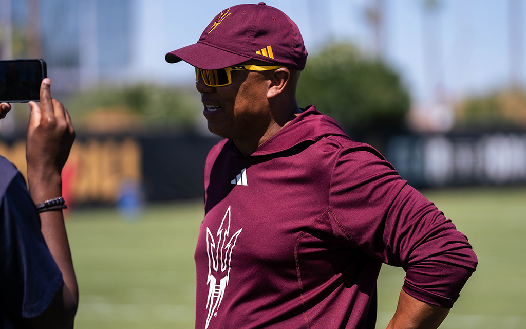 Hines Ward in a maroon hoodie and cap speaking to the media, standing on a grassy field under a clear sky.