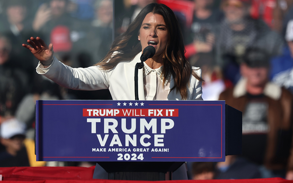 Danica Patrick speaking at a podium with a Trump/Vance sign in front of a crowd.