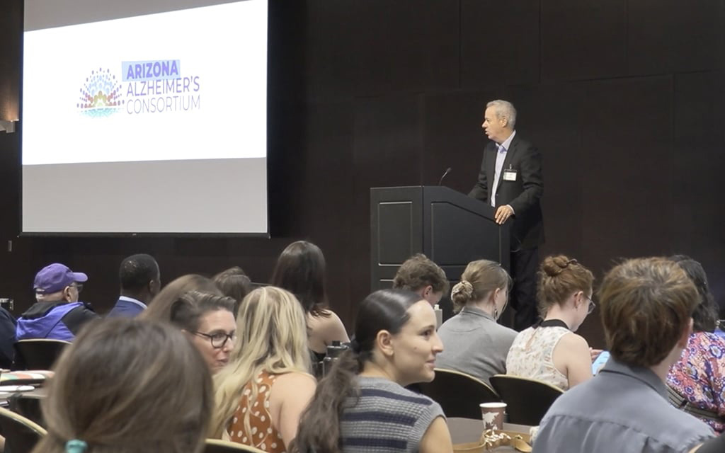 A man speaks at a podium next to a screen displaying the "Arizona Alzheimer's Consortium" logo in a conference room.