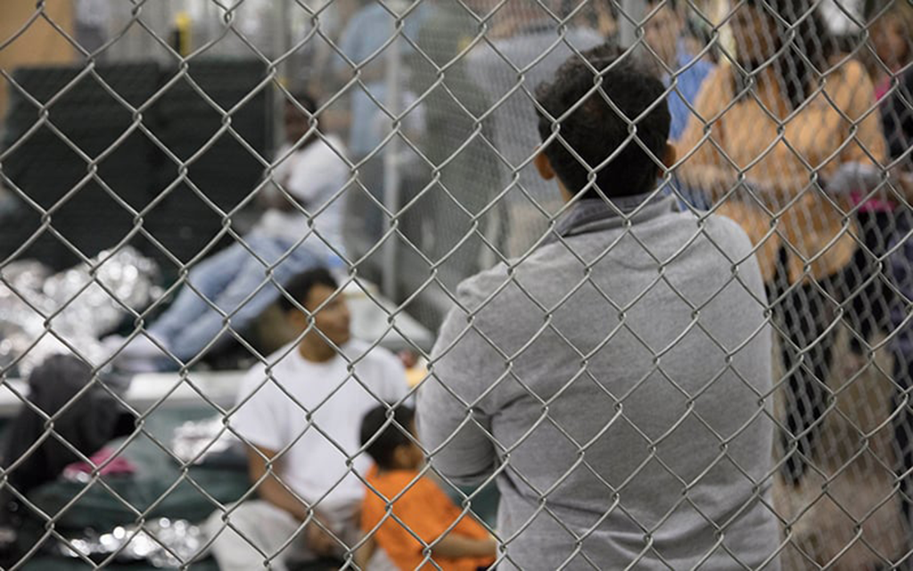 A view through a chain-link fence showing individuals inside, including a person in gray facing forward.