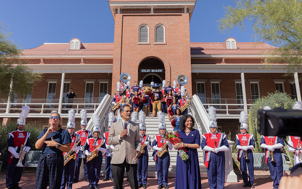 Three people stand in front of a brick building outside with a marching band behind them.