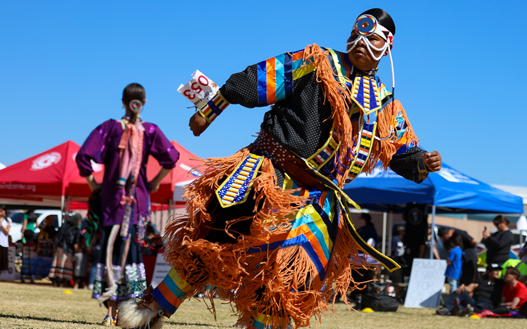 Dancer in colorful traditional attire performs at an outdoor event with tents and attendees in the background.
