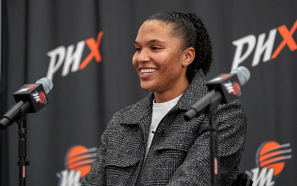 Alyssa Thomas smiling at a table with microphones, against a background with "PHX" lettering.