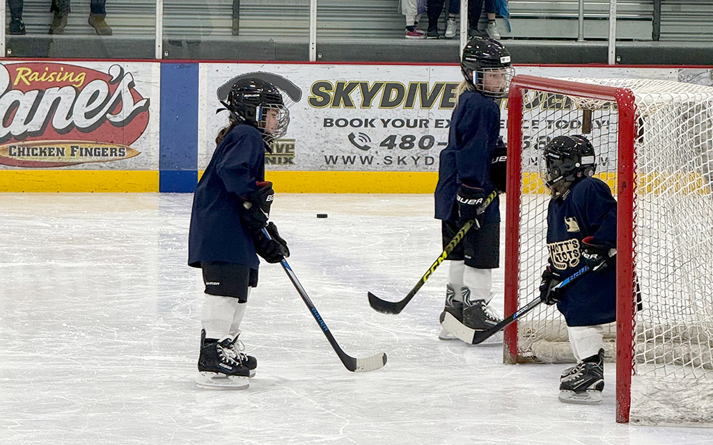 The image shows three young children at an indoor ice hockey rink, all wearing matching navy blue jerseys, black helmets, white hockey pants, and black skates. They each hold a hockey stick. One child stands to the left on the ice, facing the other two children who stand near a red and white hockey goal post.