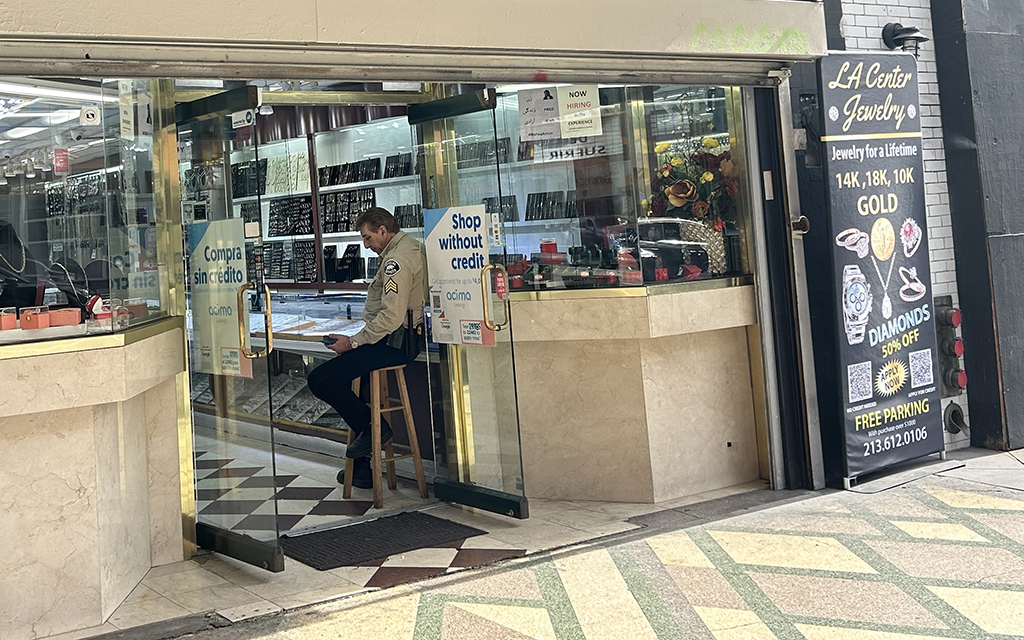 Entrance of a jewelry store with a security guard seated and promotional signs on the glass doors.