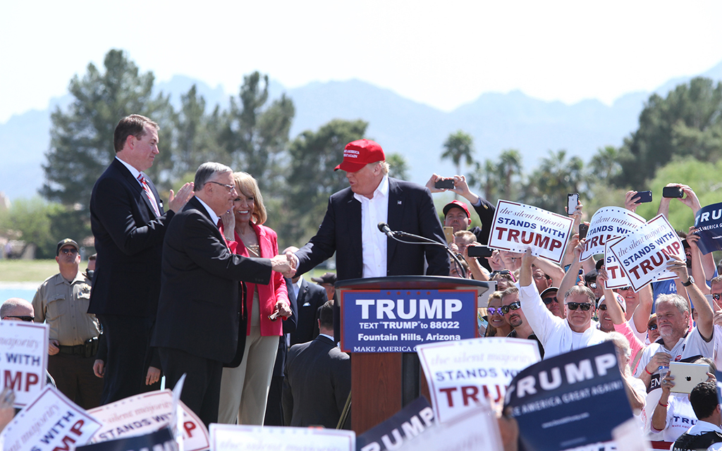President Donald Trump shakes hands with another man at a podium surrounded by people holding "TRUMP" signs. Trees and a blue sky are visible.