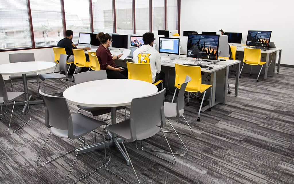 A computer lab with round tables and gray chairs in the foreground, and rows of desks with desktop computers and yellow chairs in the background.