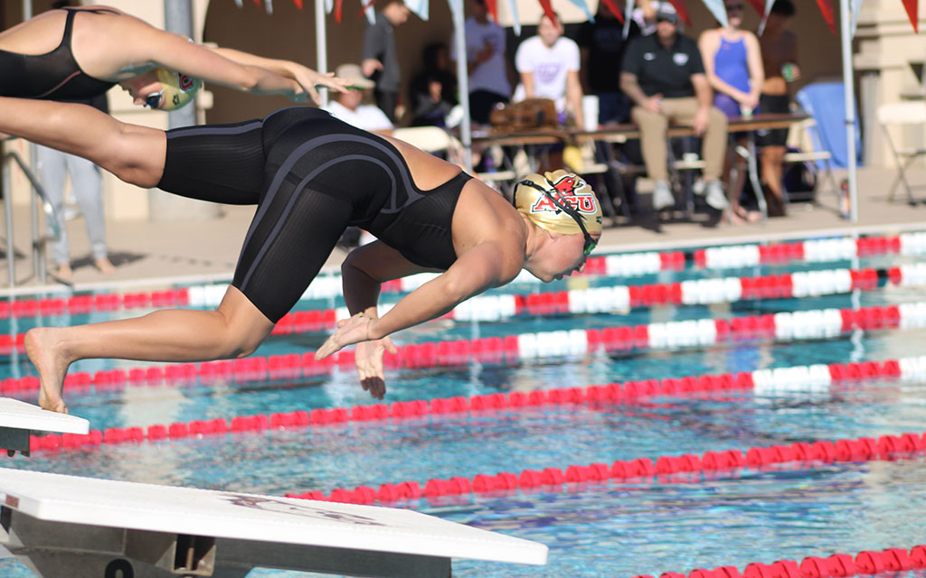 Swimmer diving into a pool during a competition, wearing a black swimsuit and beige swim cap, with spectators in the background.