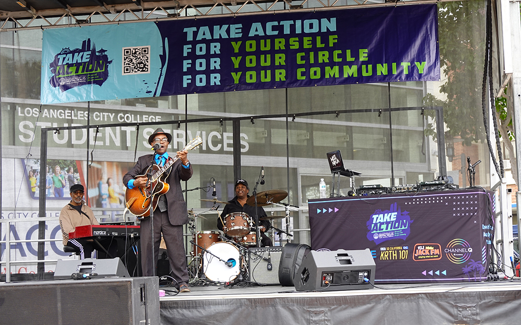 Musicians perform on stage in front of Los Angeles City College with a banner reading "TAKE ACTION FOR YOURSELF FOR YOUR CIRCLE FOR YOUR COMMUNITY."