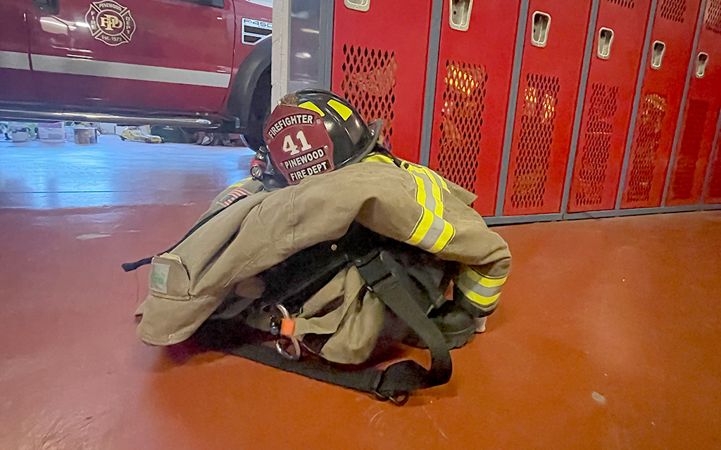 Firefighting gear, including a helmet and jacket, on a red floor with lockers and a fire truck in the background.