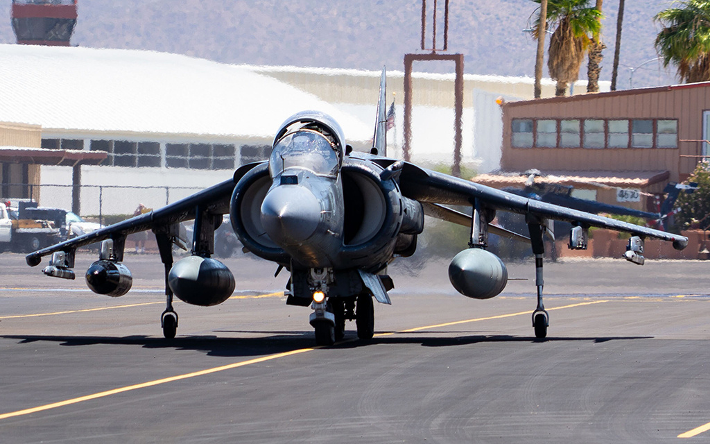 A military jet on a runway with hangars and palm trees in the background.