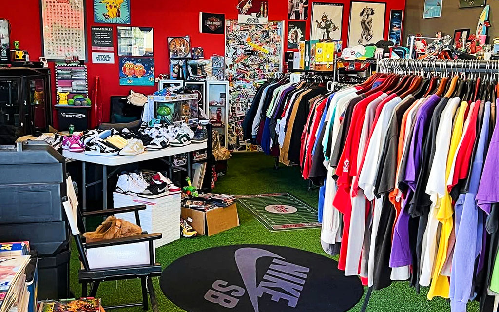 The image shows the interior of a vibrant store with a red wall adorned with various posters and artwork. To the left, there is a table filled with an assortment of sneakers in different styles and colors. Beneath the table, more shoes are visible. On the right side, a clothing rack is densely packed with colorful t-shirts hung on black hangers. The floor is covered with green artificial turf, and a black circular mat with a large "Nike SB" logo occupies the foreground. The back of the store features more posters and a cluttered display of merchandise, adding to the eclectic and busy atmosphere.