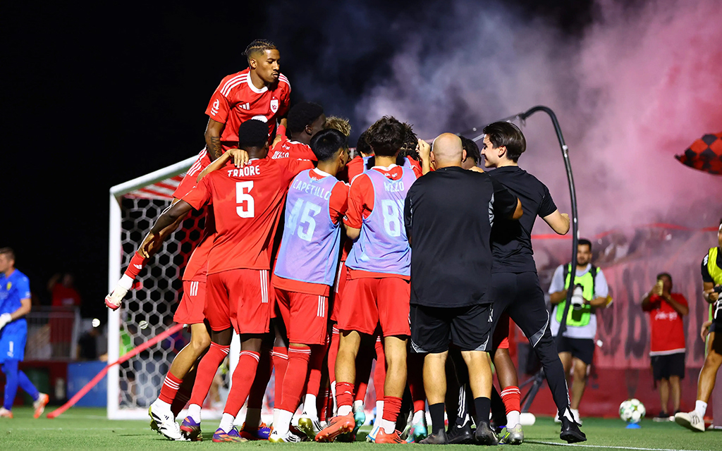 Phoenix Rising players in red uniforms celebrating near a goalpost with red smoke in the background.