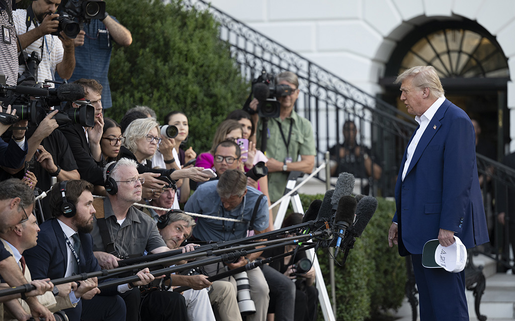 President Donald Trump is facing a group of journalists with cameras and microphones outside a large white building.