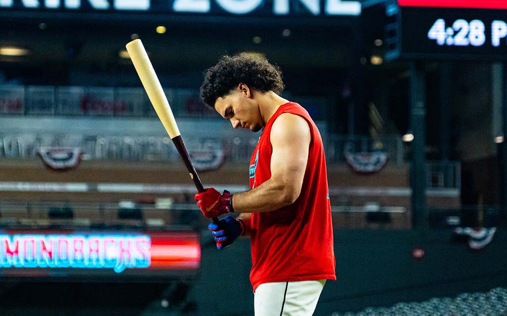 Alex Thomas in a red sleeveless shirt holds a bat in a stadium.