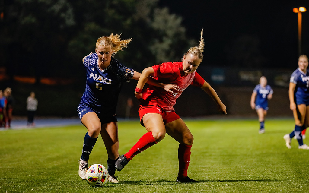 The image depicts a nighttime soccer game on a grassy field with players engaged in action. In the foreground, two female soccer players are competing for the ball. The player on the left is wearing a blue jersey with number 5 and "NAU" printed on it, complementing blue shorts and socks. Her hair is tied back, and she is intensely focused on the ball. The player on the right is dressed in red, with a red jersey and shorts, as well as red knee-high socks.