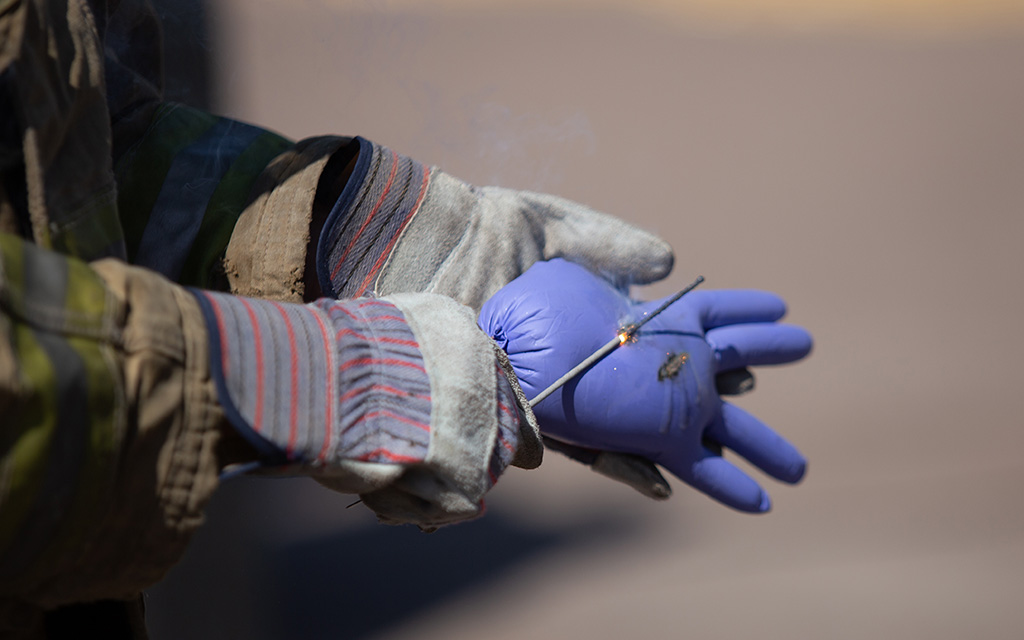 The image shows a close-up of a person wearing protective gear, including thick striped gloves and a fire-resistant jacket with reflective stripes. They are holding a burning wire in their right hand, which is gloved with a blue latex glove. A small flame or spark is visible on the wire, emitting a slight trail of smoke against a blurred background.