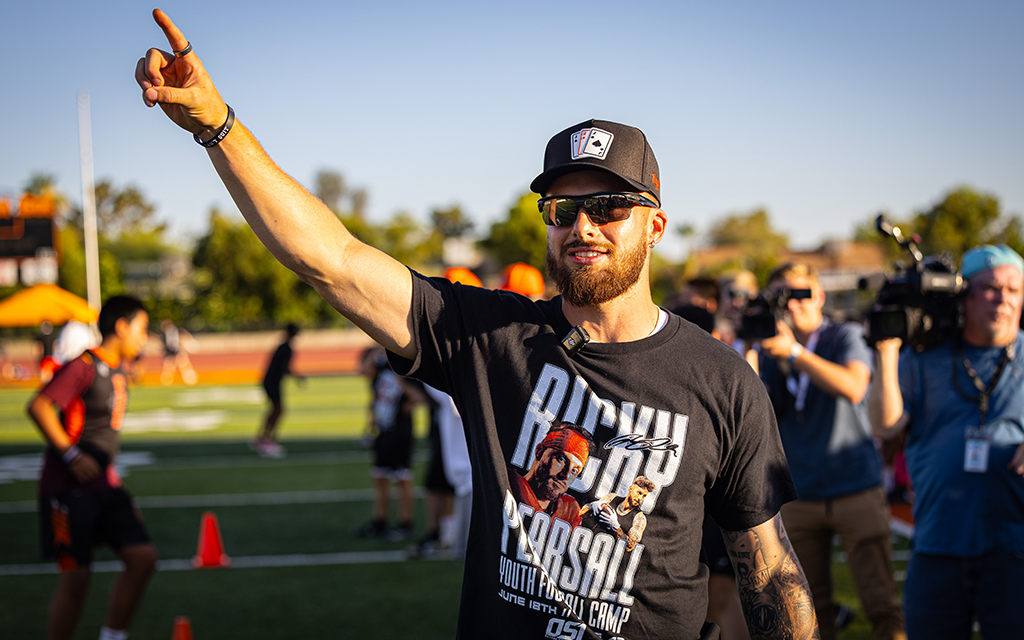 Man in a black t-shirt and sunglasses pointing on a sports field.